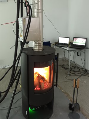 Ein Foto des Kaminofenprüfstands im Betrieb. In der Brennkammer sind einige Holzscheite und Feuer zu sehen. Vom Kamin gehen diverse Kabel und Schläuche aus. Im Hintergrund sind zwei Laptops zu erkennen.