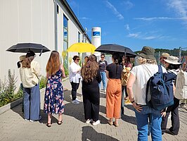 Eine Gruppe steht mit Sonnenschirmen vor einem Fabrikgebäude